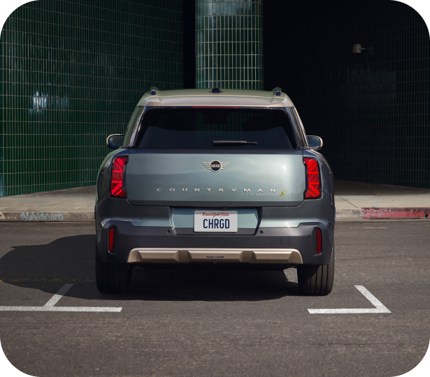 Three-quarters front view of a MINI Electric vehicle parked on a cobblestone surface in a parking space in front of a retail storefront in an urban setting.