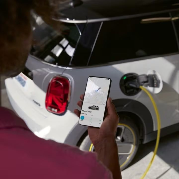 Person in pink collared shirt holding a smartphone with the MINI App displayed on its screen, plus a white MINI Electric vehicle parked in the background with a yellow charging cord plugged into it.