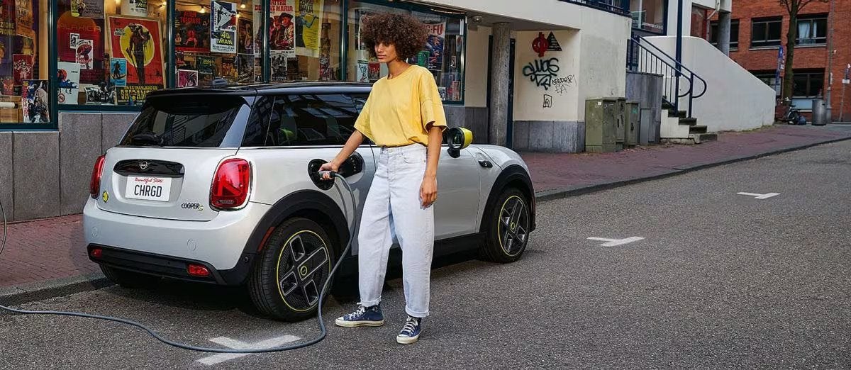 Three-quarters back view of a white MINI Cooper SE Hardtop 2 Door vehicle parked on a street, with a person in a yellow shirt plugging a charger into the vehicle plus a windowed retail storefront in the background.