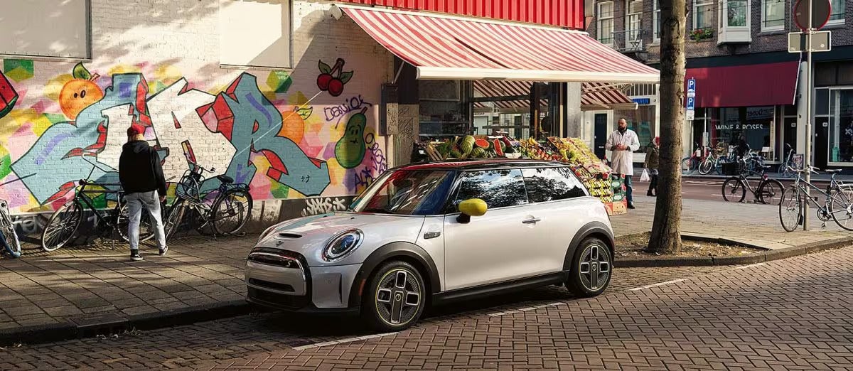 Three-quarters front view of a white MINI Cooper SE Hardtop 2 Door parked on a street in an urban setting with a graffiti wall and retail storefront in the background.