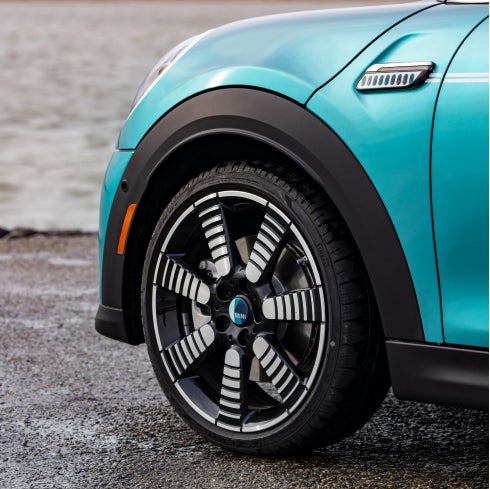 Closeup view of an 18” Pulse Spoke 2-Tone Wheel on a MINI Cooper S Convertible Seaside Edition in Caribbean Aqua, parked on a dark surface with a white wall in the background.