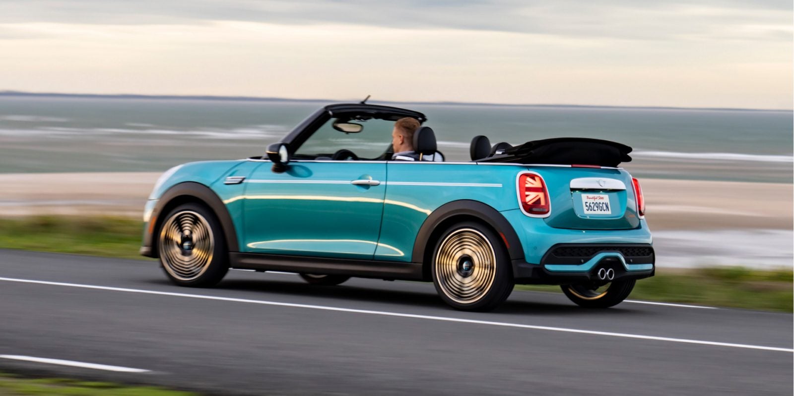 Side view of a MINI Cooper S Convertible Seaside Edition in Caribbean Aqua driving along a coastal road with a man in the driver’s seat and no one else in the vehicle, including a beach and cloudy skies in the background.